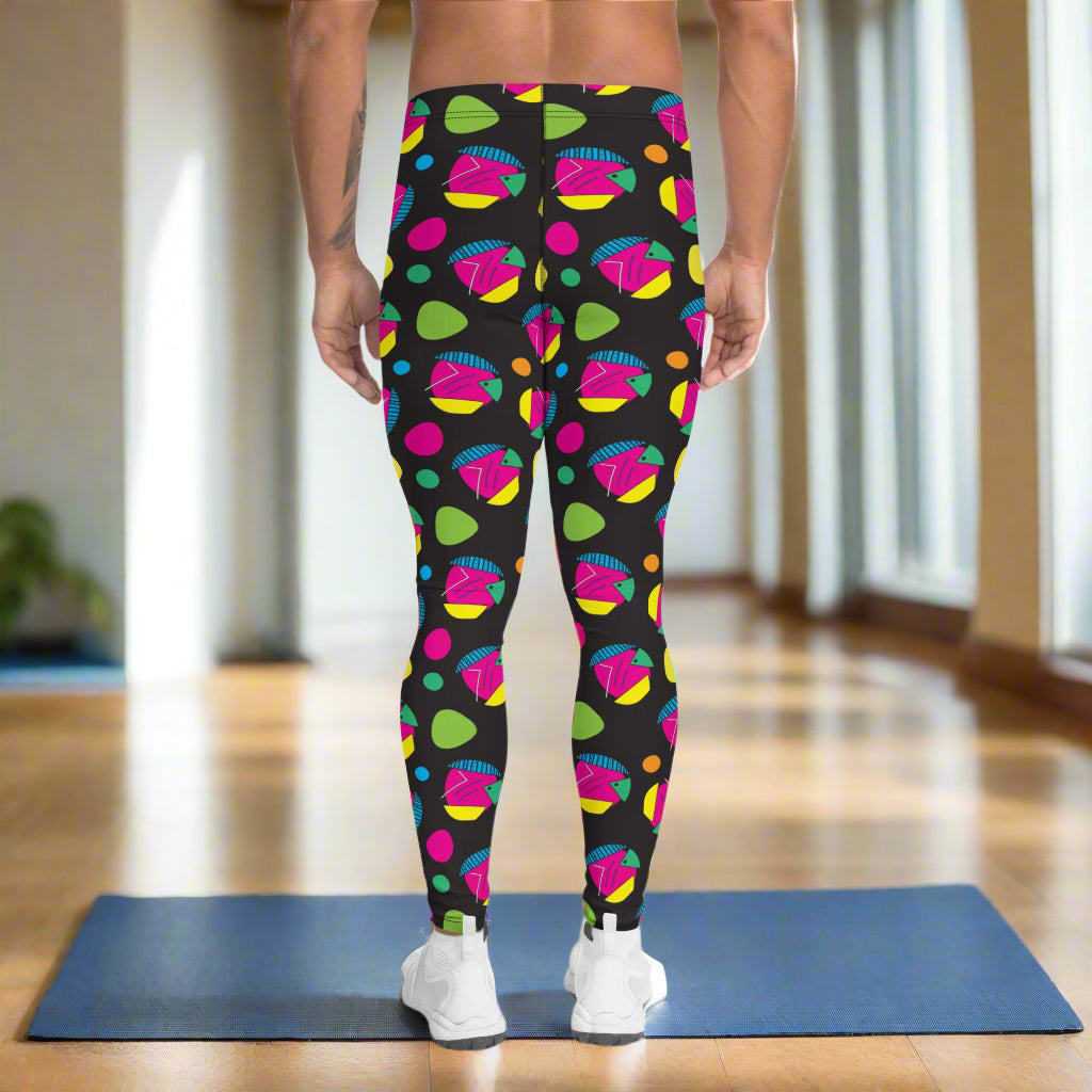 Man from the waist down wearing men's Funk Party funky festival yoga leggings standing facing away with his hands by his sides. The man is standing on a blue gym or yoga may on a wooden floor in an apartment with light streaming in from large windows to the right. The funk party pattern is an abstract pattern in vibrant pink, orange, yellow, green and blue in a spotty type arrangement on a black background