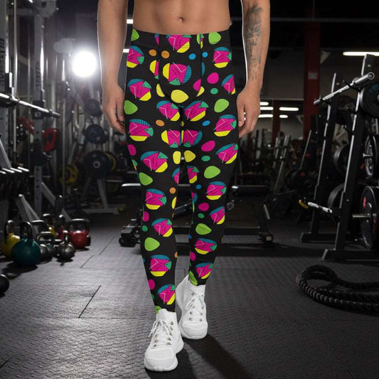 Man from the waist down wearing men's Funk Party funky festival yoga leggings standing facing forwards with his hands by his sides. The man is standing on a wooden floor in an apartment with light streaming in from a large window behind him. There is a blue gym or yoga mat and two small house plants near to the left of the window. The funk party pattern is an abstract pattern in vibrant pink, orange, yellow, green and blue in a spotty type arrangement on a black background