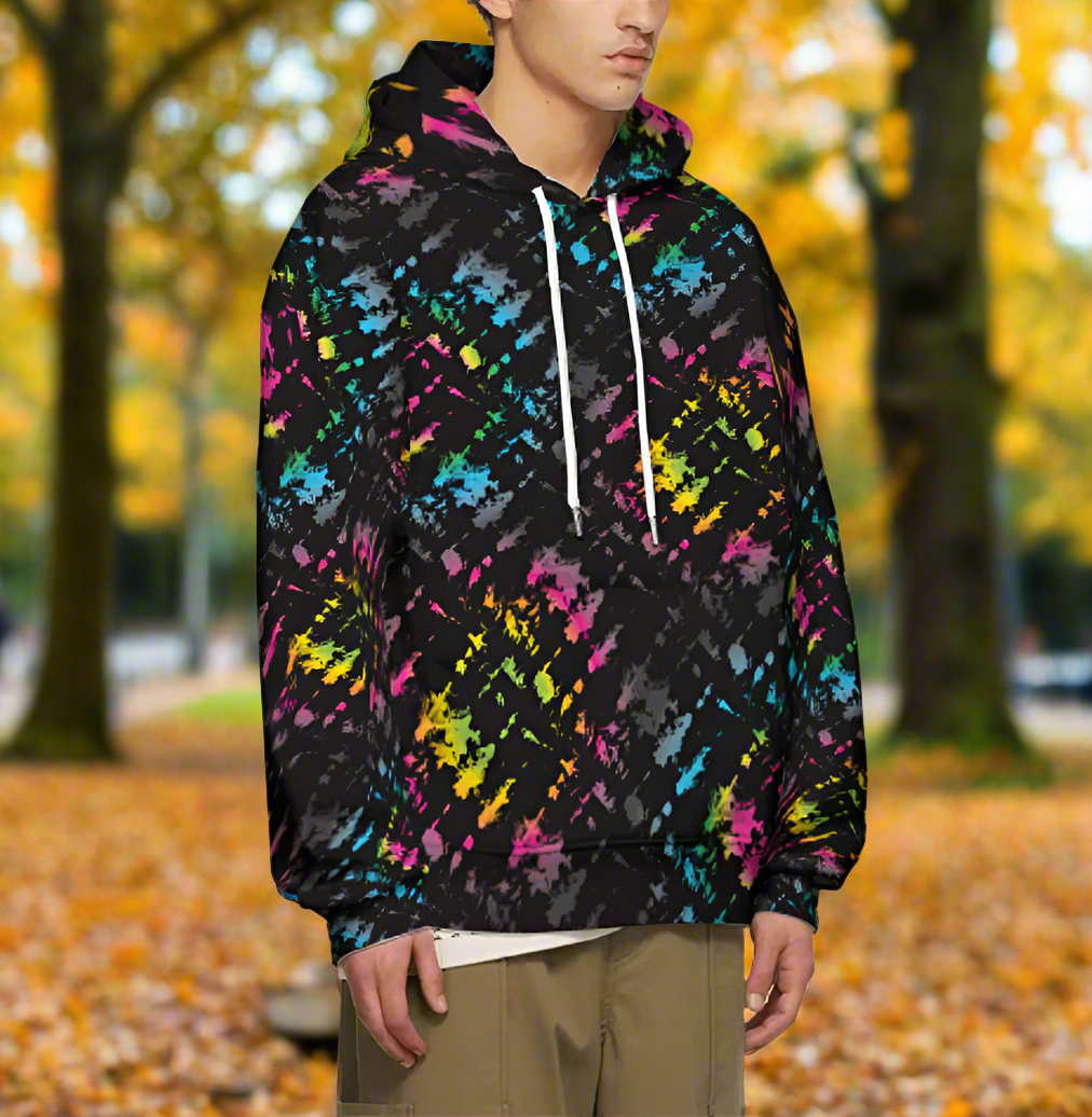 Man from the chin down to the waist, posing standing facing to the right wearing a 100% cotton vibrant Minstrel Magic tie-dye all-over print hoodie. He is standing outside amongst autumnal trees with golden leaves blanketing the ground. The hoodie is black with splashes of bright pink, orange yellow, green, blue and grey. It is a bold fun design for everyday wear.