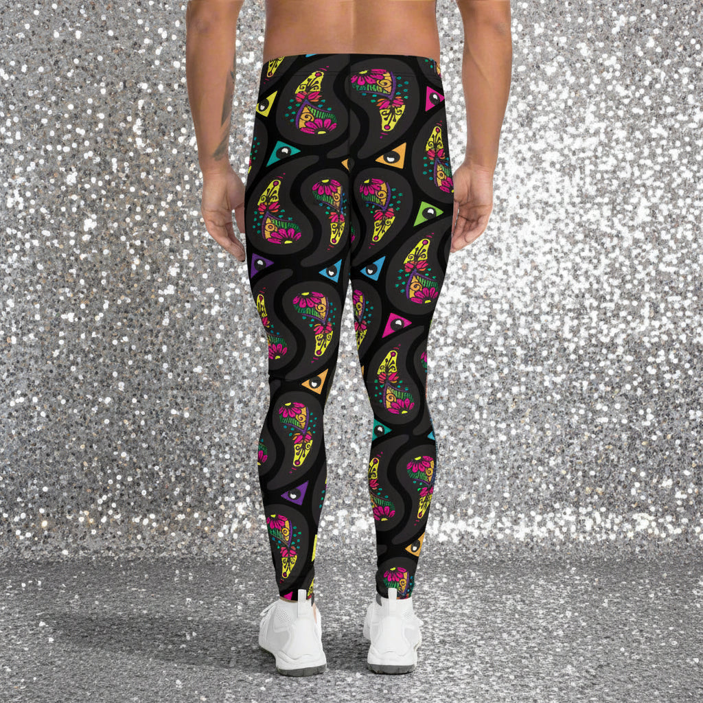 Man from the waist down wearing men's black paisley party funky festival yoga leggings standing facing away with his hands by his sides. He is wearing white trainers and is standing on a countryside green marked tennis court with trees and hedges in the background. The paisley party pattern is a colourful repeating large paisley pattern, made up of vibrant colours including pink, orange, yellow, green, blue and purple. In between the paisley print are colourful triangles in the same colours.