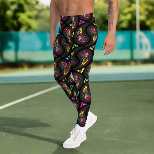 Man from the waist down wearing men's black paisley party funky festival yoga leggings standing facing to the left with his hands by his sides. He is wearing white trainers and is standing on a countryside green marked tennis court with trees in the background. The paisley party pattern is a colourful repeating large paisley pattern, made up of vibrant colours including pink, orange, yellow, green, blue and purple. In between the paisley print are colourful triangles in the same colours.