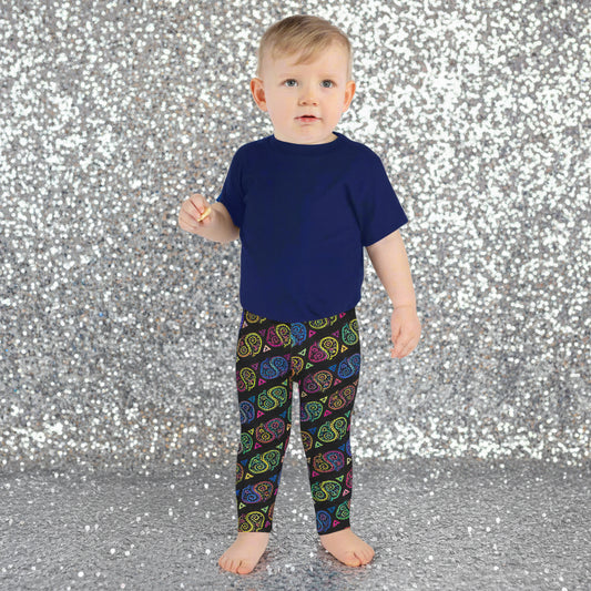A toddler wearing kids size black paisley party funky festival leggings, poses facing forwards. He is standing on a stone path that leads to a white and blue house with bright pink flowers growing in the background. The paisley party pattern is a colourful repeating large paisley pattern, made up of vibrant colours including pink, orange, yellow, green, blue and purple. In between the paisley print are colourful triangles in the same colours