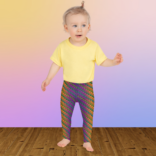 A toddler wearing kids size fire tribal frog funky festival leggings and a pale yellow t-shirt stands on a wooden floor facing forwards. She is standing in front of a pale pastel pink, purple and yellow coloured background. The fire tribal frog pattern is an abstract pattern with a blended orange, pink and purple background.