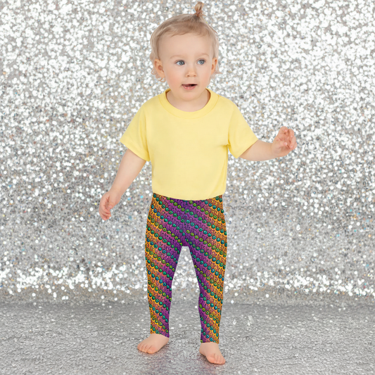 A toddler wearing kids size fire tribal frog funky festival leggings and a pale yellow t-shirt stands on a wooden floor facing forwards. She is standing in front of a pale pastel pink, purple and yellow coloured background. The fire tribal frog pattern is an abstract pattern with a blended orange, pink and purple background.