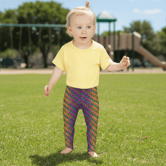 A toddler wearing kids size fire tribal frog funky festival leggings and a pale yellow t-shirt stands on a wooden floor facing forwards. She is standing in front of a pale pastel pink, purple and yellow coloured background. The fire tribal frog pattern is an abstract pattern with a blended orange, pink and purple background.