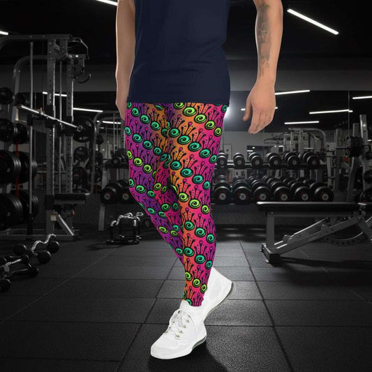 Man from the waist down wearing men's fire tribal frog funky festival yoga leggings standing facing to the left with his hands by his sides. He is wearing white trainers and is standing in a grassy festival field with other festival goers mingling in the background. It is a sunny day. The fire tribal frog pattern is a funky abstract pattern with a blended pink, purple and orange background.