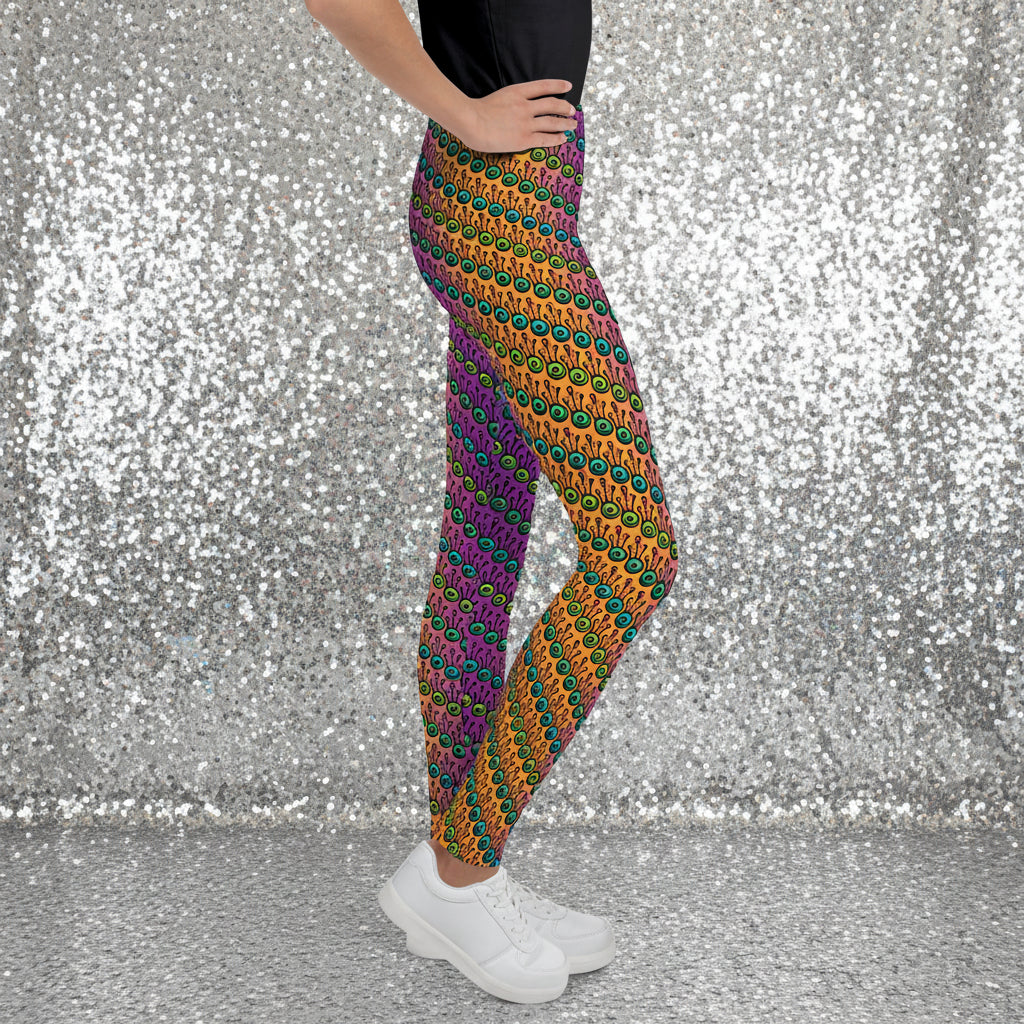 A youth pictured from the waist down wearing youth size fire tribal frog funky festival leggings standing facing to the right with both hands on their hips. They are wearing white trainers and standing on a wooden gymnasium floor with a cream wall. The fire tribal frog pattern is an abstract pattern with a blended orange, pink and purple background.