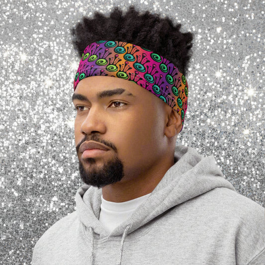 Man with short, dark, afro hair is facing to the left wearing a grey hoodie and a vibrant tribal frog neck gaiter around his head, as a head band. In the background are some fairy lights and plants. The fire tribal frog design is an abstract pattern with a blended purple, pink and orange background. The abstract detail is coloured in different shades of green.