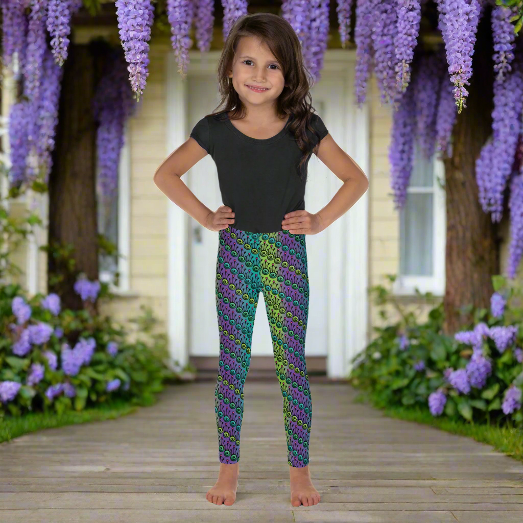 A young girl wearing kids size jungle tribal frog funky festival leggings, stands facing forwards with her hands on her hips. She is smiling and standing in front of a white door to a country house, with blooming wisteria flowers draping over the house. The jungle tribal frog pattern is an abstract pattern with a blended green and purple background.