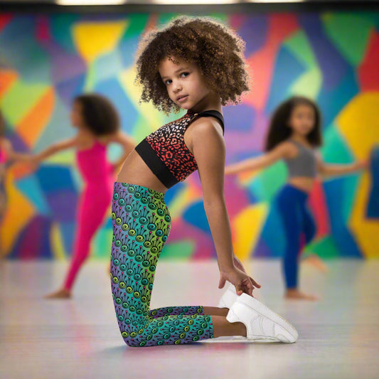 A young girl poses wearing kids size jungle tribal frog funky festival leggings. She is in a brightly coloured dance studio with other young dancers in the background. The back wall is painted in vibrant multicoloured abstract shapes. The jungle tribal frog pattern is an abstract pattern with a blended green and purple background.