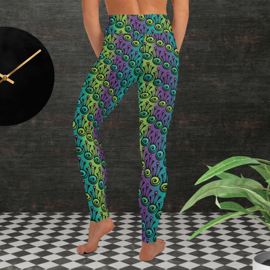 Woman from the waist down wearing high-waist Tribal Frog funky festival yoga leggings standing facing backwards with her hands down to her front. The woman is standing on a black and white tiled floor in front of a textured grey background with a black and gold wall clock to the left and a partially visible leafy plant on the floor in the bottom right. The jungle tribal frog pattern is an abstract pattern with a blended green and purple background.