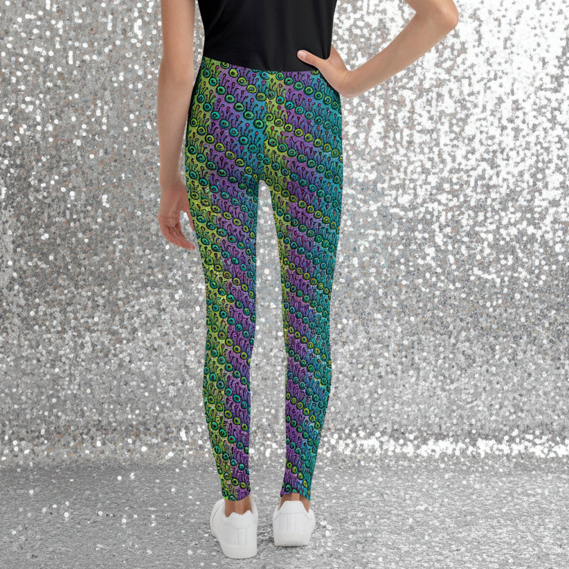 A youth pictured from the waist down wearing youth size jungle tribal frog funky festival leggings standing facing away with their right hand on their hip. They are wearing white trainers and standing on a wooden dance studio floor against a light grey wall. The jungle tribal frog pattern is an abstract pattern with a blended green and purple background.