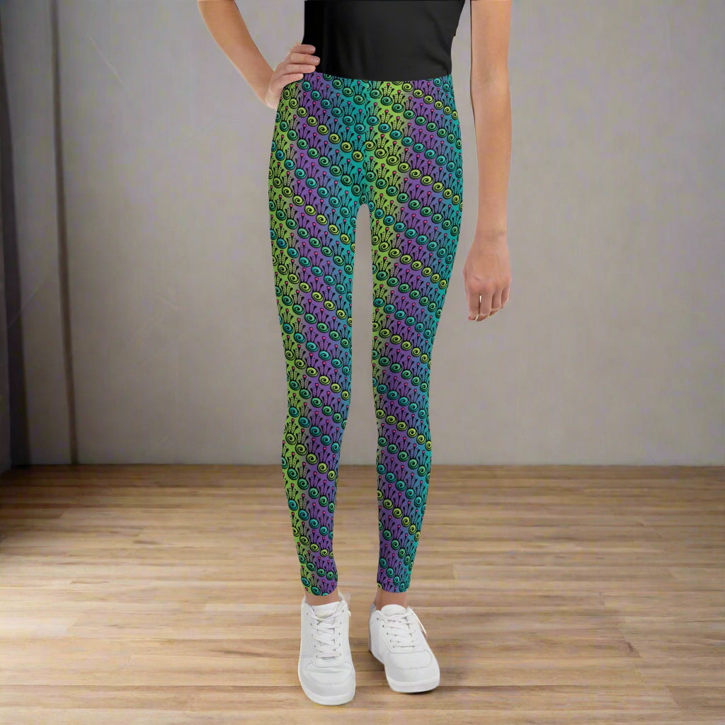 A youth pictured from the waist down wearing youth size jungle tribal frog funky festival leggings standing facing forwards with their right hand on their hip. They are wearing white trainers and standing on a wooden dance studio floor against a light grey wall. The jungle tribal frog pattern is an abstract pattern with a blended green and purple background.