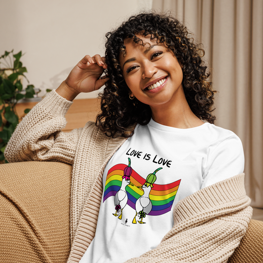 Woman sitting relaxing and smiling at the camera. She is in a living area on comfy seating, wearing a fitted white Sassy Seagull Love Is Love LGBTQ+ Pride T-Shirt and a cream knitted cardigan. The t-shirt is white with short sleeves. The graphic shows two cartoon style seagulls wearing colourful hats walking away in front of an LGBTQIA pride flag, underneath the wording Love Is Love. It is a bold playful design for everyday wear.