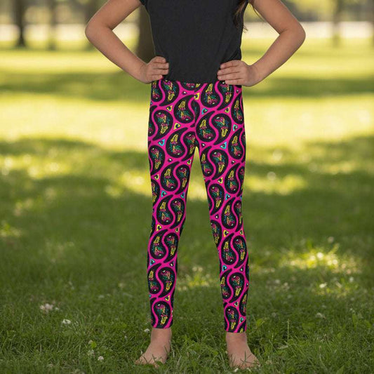 A young girl poses facing to the front outside an ornate orange and turquoise moroccan-style building with an arch and tiled floor. She is wearing pink paisley party funky festival leggings, and a black t-shirt. The paisley party pattern is a colourful repeating large paisley pattern, made up of vibrant colours including pink, orange, yellow, green, blue and purple. In between the paisley print are colourful triangles in the same colours. The pattern background is hot pink