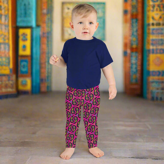 A toddler poses facing forward inside an ornate orange and turquoise moroccan-style building with a tiled floor. He is wearing pink paisley party funky festival leggings, and a navy t-shirt. The paisley party pattern is a colourful repeating large paisley pattern, made up of vibrant colours including pink, orange, yellow, green, blue and purple. In between the paisley print are colourful triangles in the same colours. The pattern background is hot pink