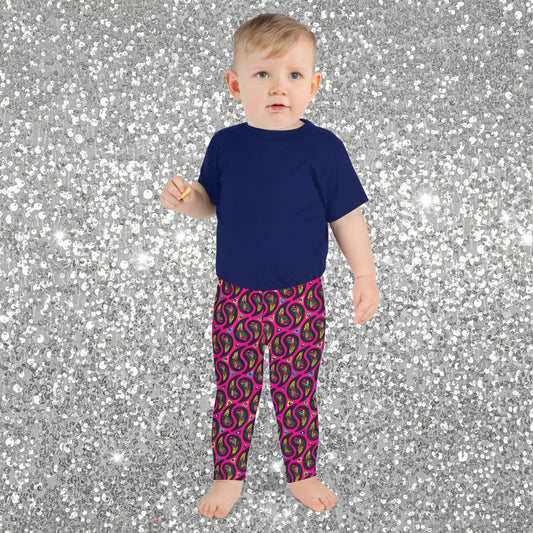 A toddler poses facing forward inside an ornate orange and turquoise moroccan-style building with a tiled floor. He is wearing pink paisley party funky festival leggings, and a navy t-shirt. The paisley party pattern is a colourful repeating large paisley pattern, made up of vibrant colours including pink, orange, yellow, green, blue and purple. In between the paisley print are colourful triangles in the same colours. The pattern background is hot pink