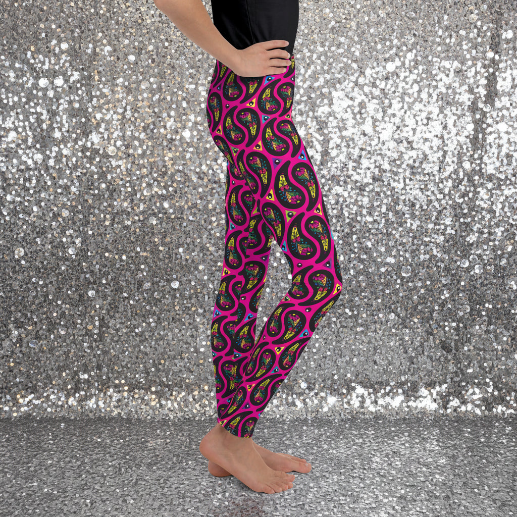 A young person from the waist down poses facing to the right inside an ornate gold and turquoise moroccan-style building with a tiled floor. They are wearing pink paisley party funky festival leggings. The paisley party pattern is a colourful repeating large paisley pattern, made up of vibrant colours including pink, orange, yellow, green, blue and purple. In between the paisley print are colourful triangles in the same colours. The pattern background is hot pink