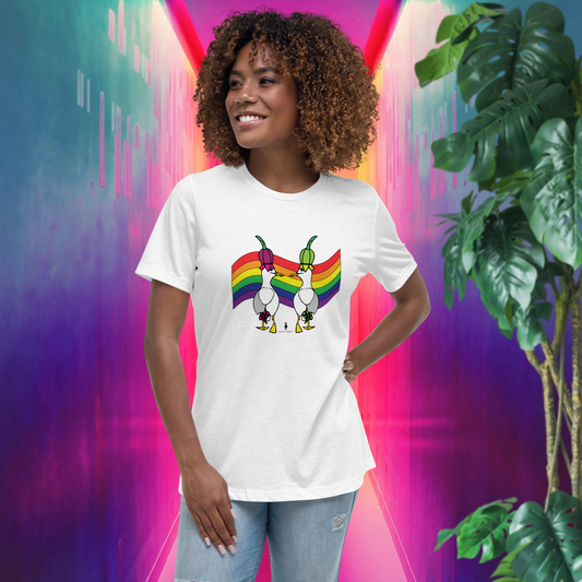 Woman standing facing forwards and looking to the left, wearing a fitted white Sassy Pride Flag LGBTQ+ Pride T-Shirt and pale stonewash blue jeans. She is standing in a rainbow coloured corridor in pinks, purples, blues and oranges with a tall green leafy plant to the front right. The t-shirt is white with short sleeves. The graphic shows two cartoon style seagulls wearing colourful hats walking away in front of an LGBTQIA pride flag. It is a bold playful design for everyday wear
