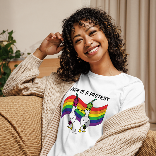 Woman sitting relaxing and smiling at the camera. She is in a living area on comfy seating, wearing a fitted white Sassy Seagull Pride is a Protest LGBTQ+ Pride T-Shirt and a cream knitted cardigan. The t-shirt is white with short sleeves. The graphic shows two cartoon style seagulls wearing colourful hats walking away in front of an LGBTQIA pride flag, underneath the wording Pride is a Protest. It is a bold playful design for everyday wear