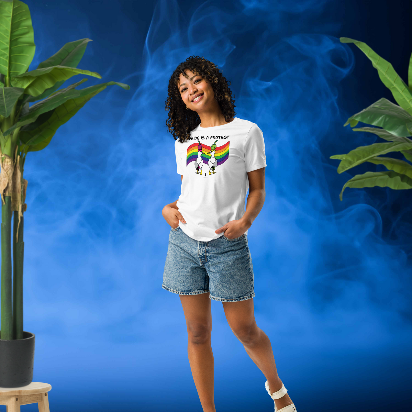 Woman posing casually facing forwards, wearing a fitted white Sassy Seagull Pride is a Protest LGBTQ+ Pride T-Shirt and stonewash blue jean shorts. She is standing in front of a smoky blue background with plants either side. The t-shirt is white with short sleeves. The graphic shows two cartoon style seagulls wearing colourful hats walking away in front of an LGBTQIA pride flag, underneath the wording Pride is a Protest. It is a bold playful design for everyday wear