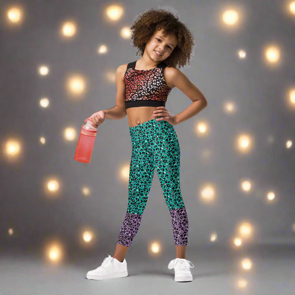 A young girl poses facing forwards while holding a water bottle. She is wearing kids size purple and green leopard glamour funky festival leggings. She is smiling and standing in front of a grey backdrop with white light orbs from fair lights around her. This design features an animal print pattern in black, grey and white, printed at a medium scale. The top 3/4 background is mint green with the bottom 1/4 purple
