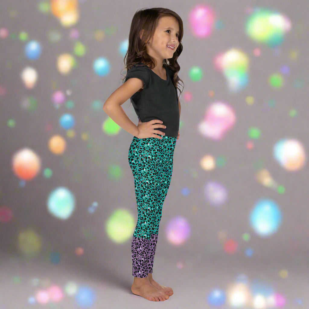 A young girl wearing kids size purple and green leopard glamour funky festival leggings stands facing to the right. She is smiling and has her hands on her hips. She is standing in front of a grey backdrop surrounded by multicoloured light orbs. This design features an animal print pattern in black, grey and white, printed at a medium scale. The top 3/4 background is mint green with the bottom 1/4 purple