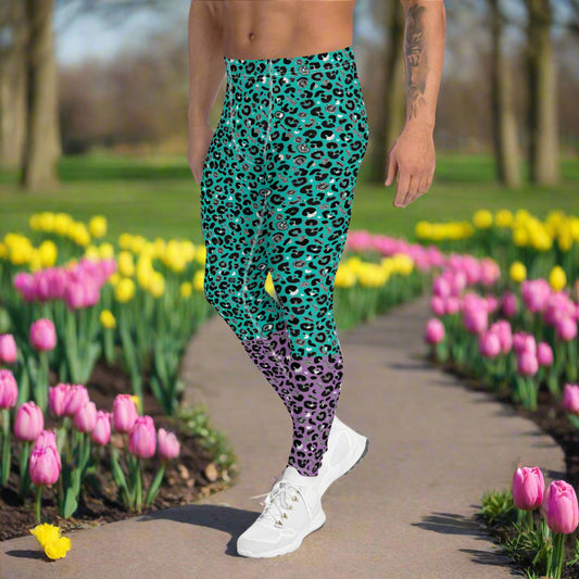 Man from the waist down wearing mens Purple and Green Leopard Glamour funky festival yoga leggings standing facing to the left with his hands by his sides. The man is standing in a park in spring with pink and yellow tulips flowering and trees in the background. This design features an animal print pattern in black, grey and white, printed at a medium scale. The top 3/4 background is mint green with the bottom 1/4 purple