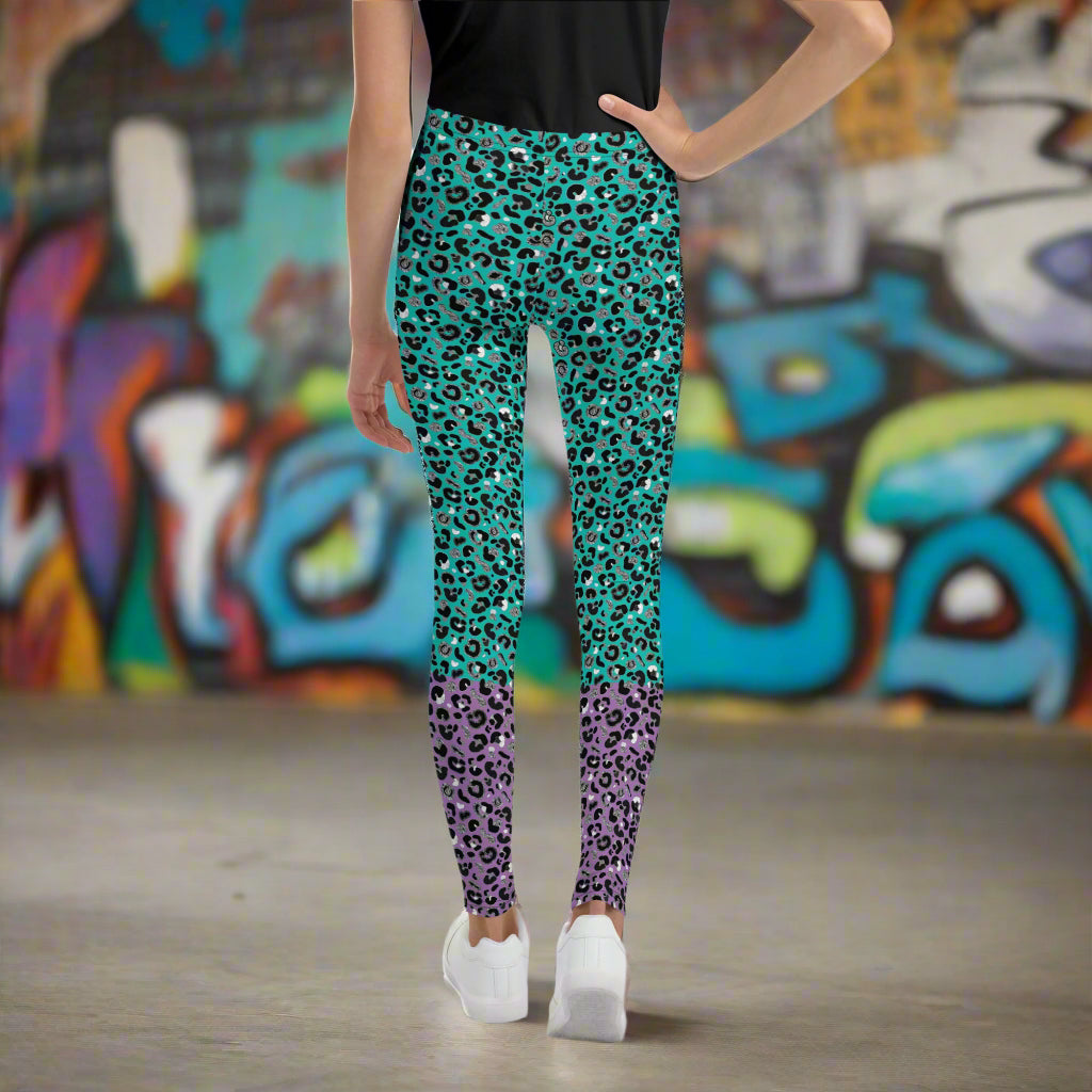 A young person from the waist down wearing youth size purple and green leopard glamour funky festival leggings stands facing away. They are standing on a concrete floor in front of a graffiti covered wall. This design features an animal print pattern in black, grey and white, printed at a medium scale. The top 3/4 background is mint green with the bottom 1/4 purple