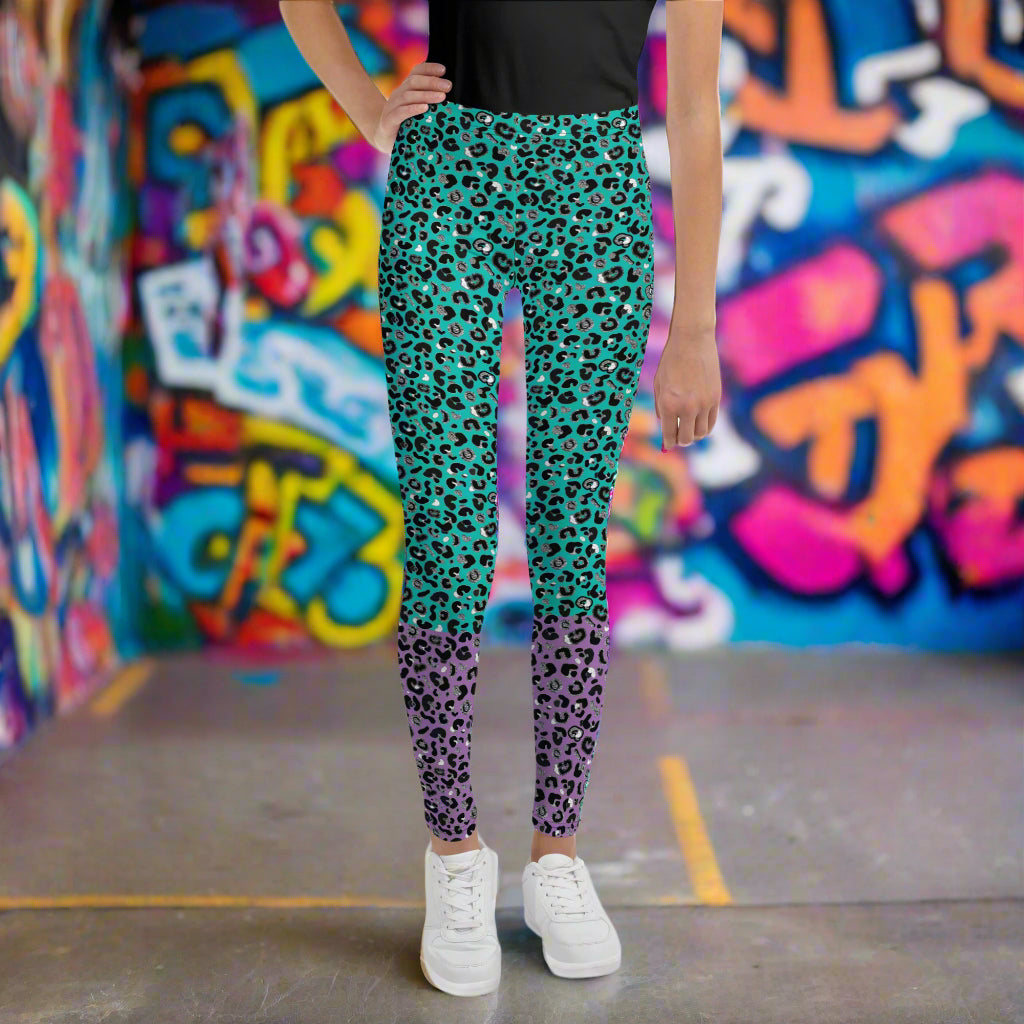 A young person from the waist down wearing youth size purple and green leopard glamour funky festival leggings stands facing forwards. They are standing on a concrete floor in front of a graffiti covered wall. This design features an animal print pattern in black, grey and white, printed at a medium scale. The top 3/4 background is mint green with the bottom 1/4 purple