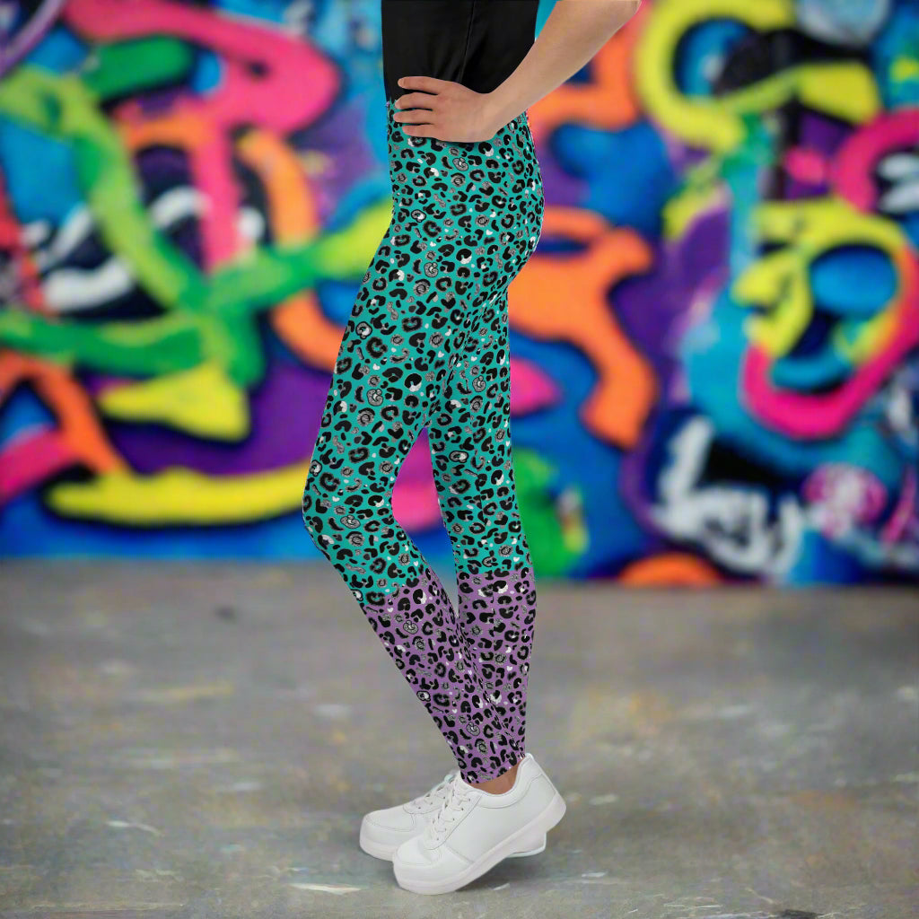 A young person from the waist down wearing youth size purple and green leopard glamour funky festival leggings stands facing to the left. They are standing on a concrete floor in front of a graffiti covered wall. This design features an animal print pattern in black, grey and white, printed at a medium scale. The top 3/4 background is mint green with the bottom 1/4 purple