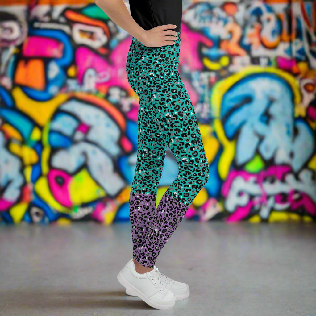 A young person from the waist down wearing youth size purple and green leopard glamour funky festival leggings stands facing to the right. They are standing on a concrete floor in front of a graffiti covered wall. This design features an animal print pattern in black, grey and white, printed at a medium scale. The top 3/4 background is mint green with the bottom 1/4 purple