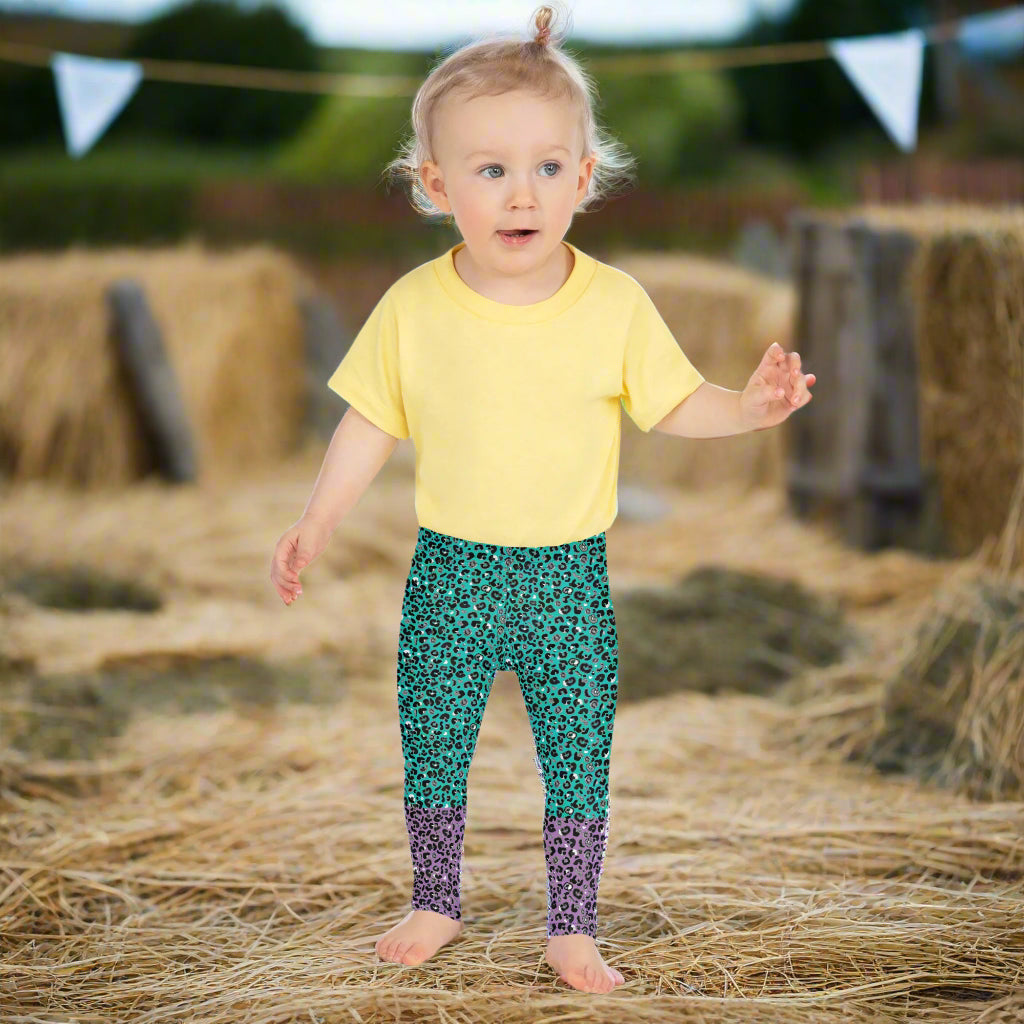 A toddler wearing kids size purple and green leopard glamour funky festival leggings stands facing forwards. She is standing amongst bales of hay with fields in the background and bunting decoration. This design features an animal print pattern in black, grey and white, printed at a medium scale. The top 3/4 background is mint green with the bottom 1/4 purple