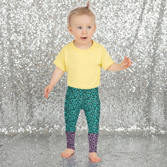 A toddler wearing kids size purple and green leopard glamour funky festival leggings stands facing forwards. She is standing amongst bales of hay with fields in the background and bunting decoration. This design features an animal print pattern in black, grey and white, printed at a medium scale. The top 3/4 background is mint green with the bottom 1/4 purple