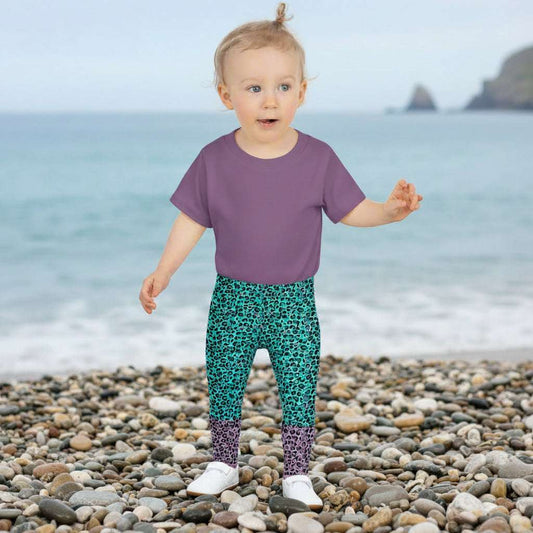 A toddler wearing kids size purple and green leopard glamour funky festival leggings stands facing forwards. She is standing amongst bales of hay with fields in the background and bunting decoration. This design features an animal print pattern in black, grey and white, printed at a medium scale. The top 3/4 background is mint green with the bottom 1/4 purple