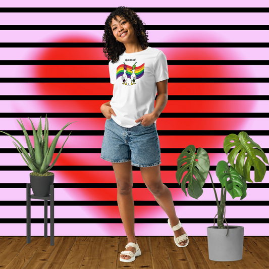 Woman posing casually facing forwards, wearing a fitted white Sassy Seagull Queer AF LGBTQ+ Pride T-Shirt and stonewash blue jean shorts. She is standing in front of a pink background with horizontal black stripes and a large, soft-focus red heart with plants to either side. The t-shirt is white with short sleeves. The graphic shows two cartoon style seagulls wearing colourful hats walking away in front of an LGBTQIA pride flag, underneath the wording Queer AF. It is a bold playful design for everyday wear
