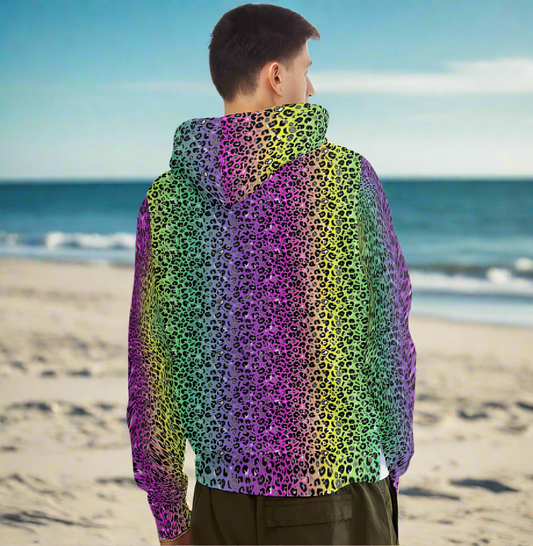 Man from the head down to the waist, posing standing facing faway wearing a 100% cotton vibrant rainbow leopard glamour all-over print animal print hoodie. He is standing on a golden sandy beach looking out towards the blue sea. The hoodie has a blended rainbow colour background from pink through to purple. The blend is a vertical stripe. The leopard print pattern is medium size in black, grey and white and sits on top of the rainbow background. It is a bold fun design for everyday wear.
