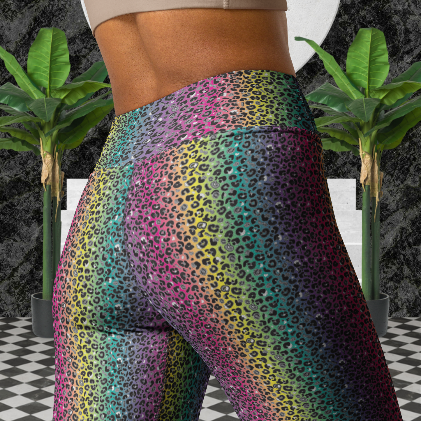 Close up of a woman from the upper back down to her thighs wearing high-waist Rainbow Leopard Glamour funky festival yoga leggings standing facing backwards. The woman is standing on a black and white tiled floor in front of some white steps and a black and grey marble background. There is a leafy plant on the floor at each side of the steps. The rainbow leopard glamour design is an animal print pattern in black, grey and white, printed at a small scale against a blended rainbow background