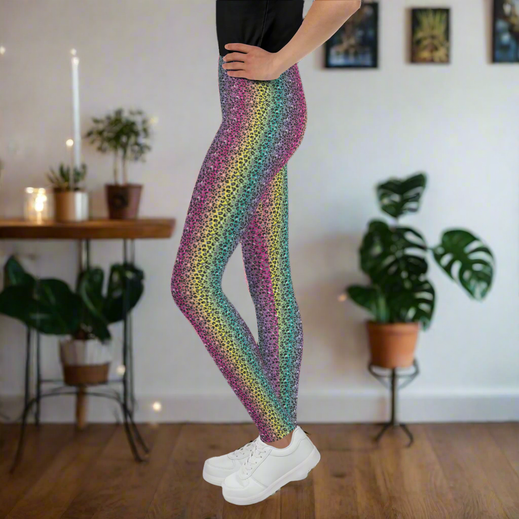 A young person pictured from the waist down wearing rainbow leopard glamour leggings and white trainers. They are facing to theft, standing on a wooden floor in a rustic home with plants and fairy lights in the background. The rainbow leopard glamour design is an animal print pattern in black, grey and white, printed at a small scale against a blended rainbow background
