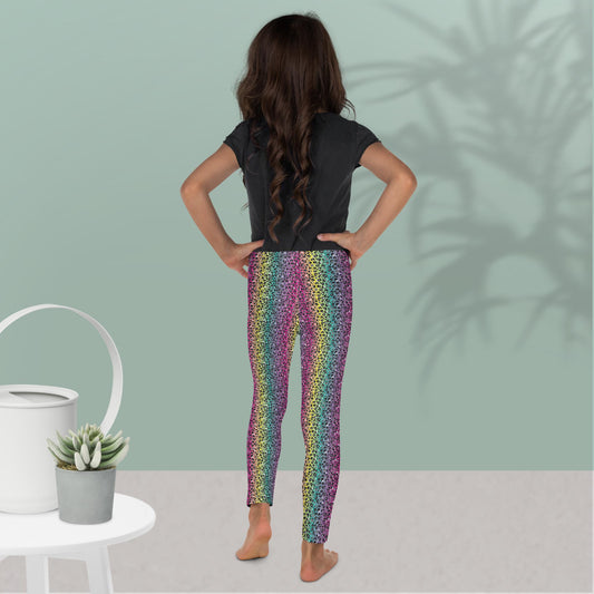 A young girl wearing kids size rainbow leopard glamour funky festival leggings and a black t-shirt, stands facing away with her hands on her hips. She is standing in front of a pastel duck egg wall with a shadow of a large spidery plant behind her. In the front left is a small white table with a small plant and white watering can on top. The rainbow leopard glamour design is an animal print pattern in black, grey and white, printed at a small scale against a blended rainbow background