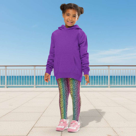A young girl wearing kids size rainbow leopard glamour funky festival leggings and an oversized grey hoodie, stands facing forwards. She is smiling and standing in front of a pastel duck egg wall with a shadow of a large spidery plant behind her. In the front left is a small white table with a small plant and white watering can on top. The rainbow leopard glamour design is an animal print pattern in black, grey and white, printed at a small scale against a blended rainbow background