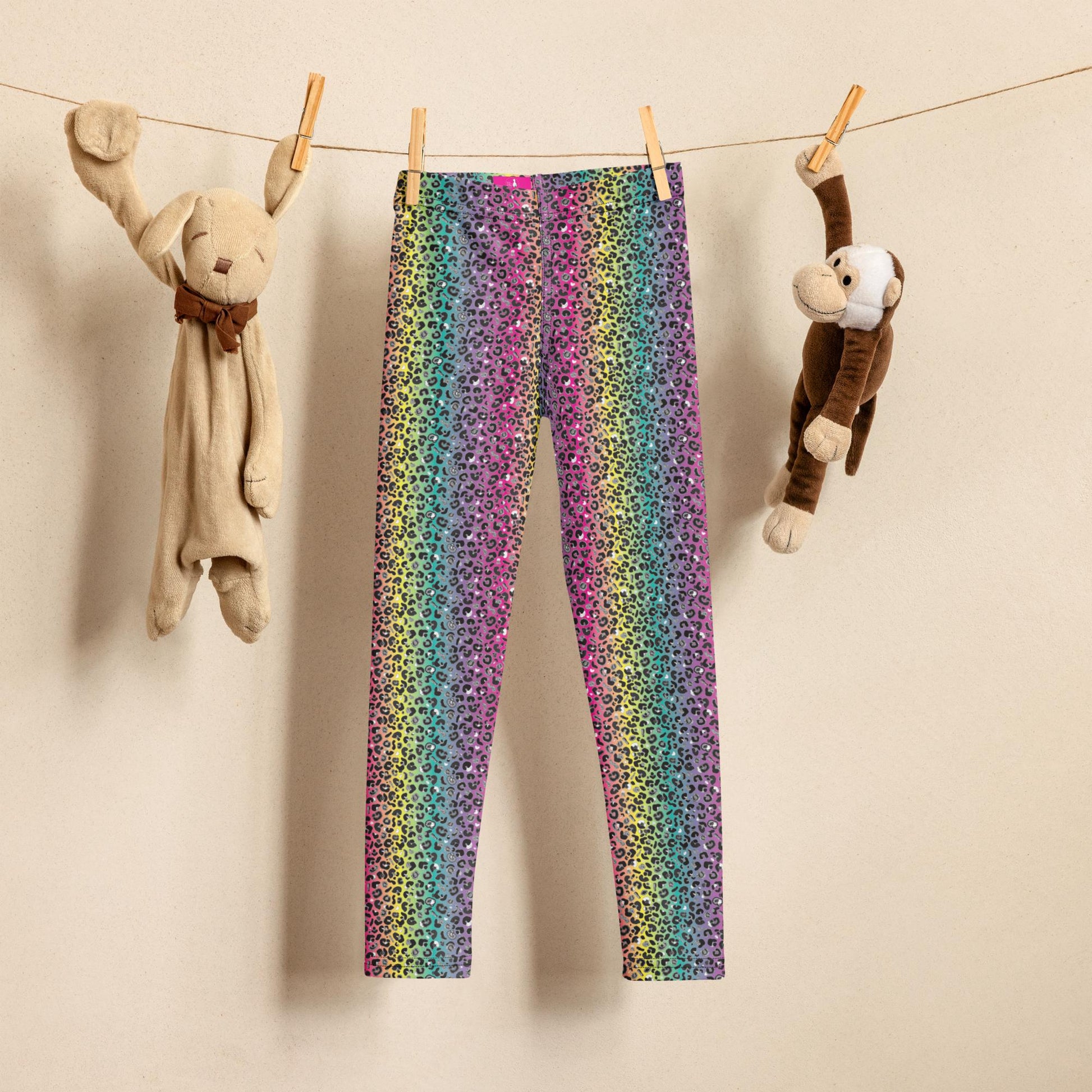 A brown string washing line hangs from the top left to the top right of the image against a pale creamy-beige background. On it, pegged with wooden pegs, hangs from left to right a comforter teddy, a pair of kids rainbow leopard glamour leggings and a small monkey teddy. The rainbow leopard glamour design is an animal print pattern in black, grey and white, printed at a small scale against a blended rainbow background