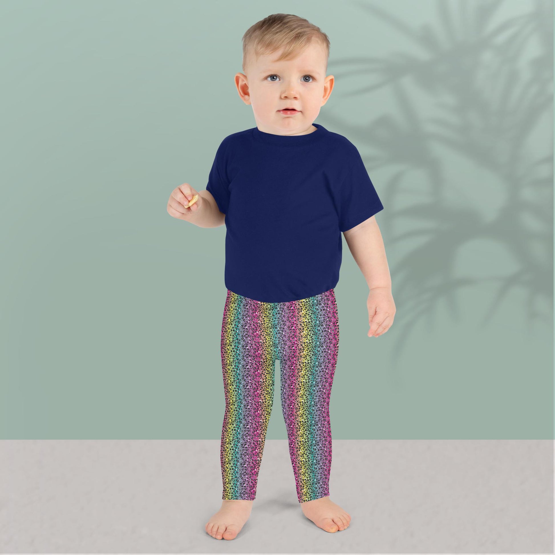 A toddler wearing kids size rainbow leopard glamour funky festival leggings and a navy blue t-shirt, stands facing forwards. He is standing in front of a pastel duck egg wall with a shadow of a large spidery plant behind him. The rainbow leopard glamour design is an animal print pattern in black, grey and white, printed at a small scale against a blended rainbow background