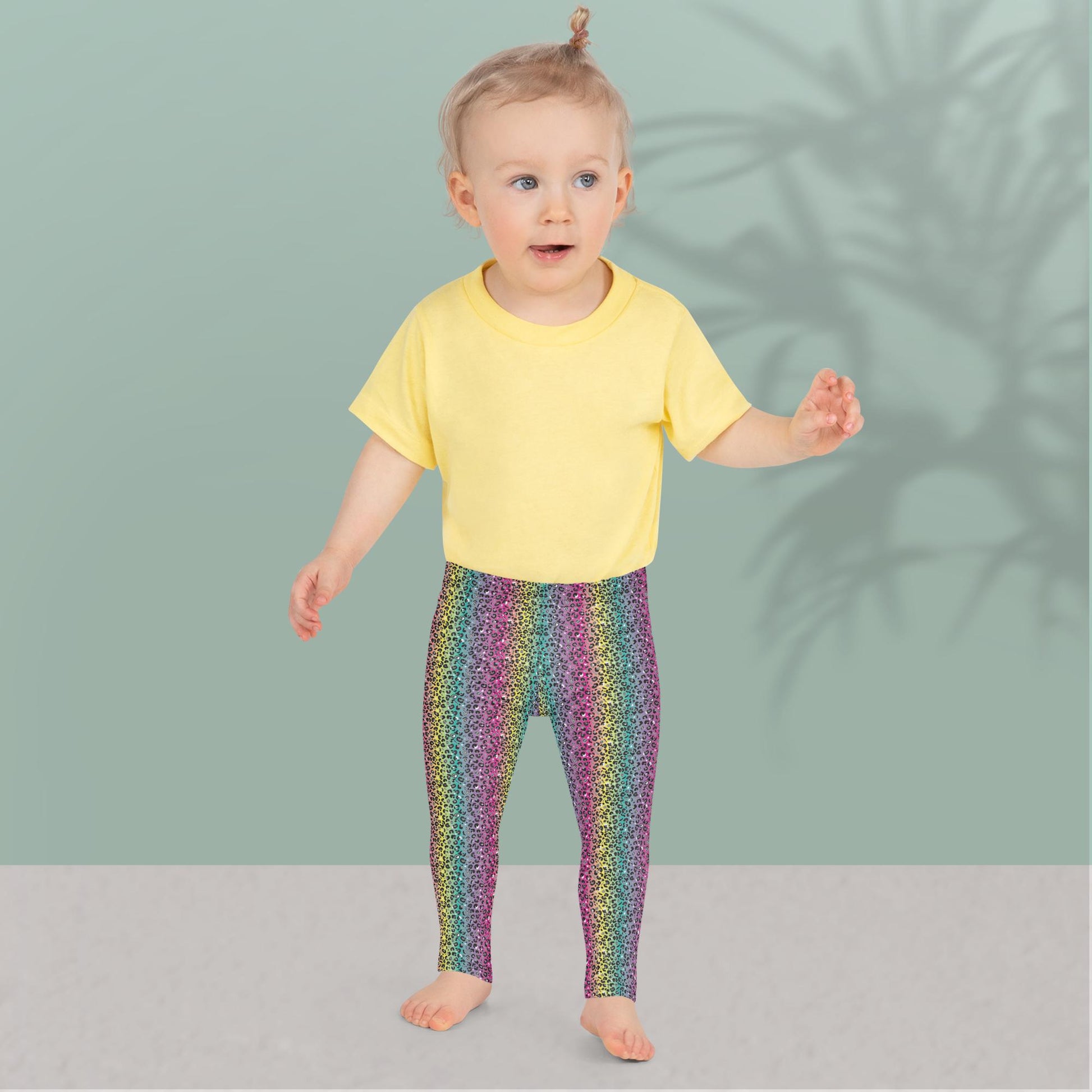 A toddler wearing kids size rainbow leopard glamour funky festival leggings and a pale yellow t-shirt, stands facing forwards. She is standing in front of a pastel duck egg wall with a shadow of a large spidery plant behind her. The rainbow leopard glamour design is an animal print pattern in black, grey and white, printed at a small scale against a blended rainbow background