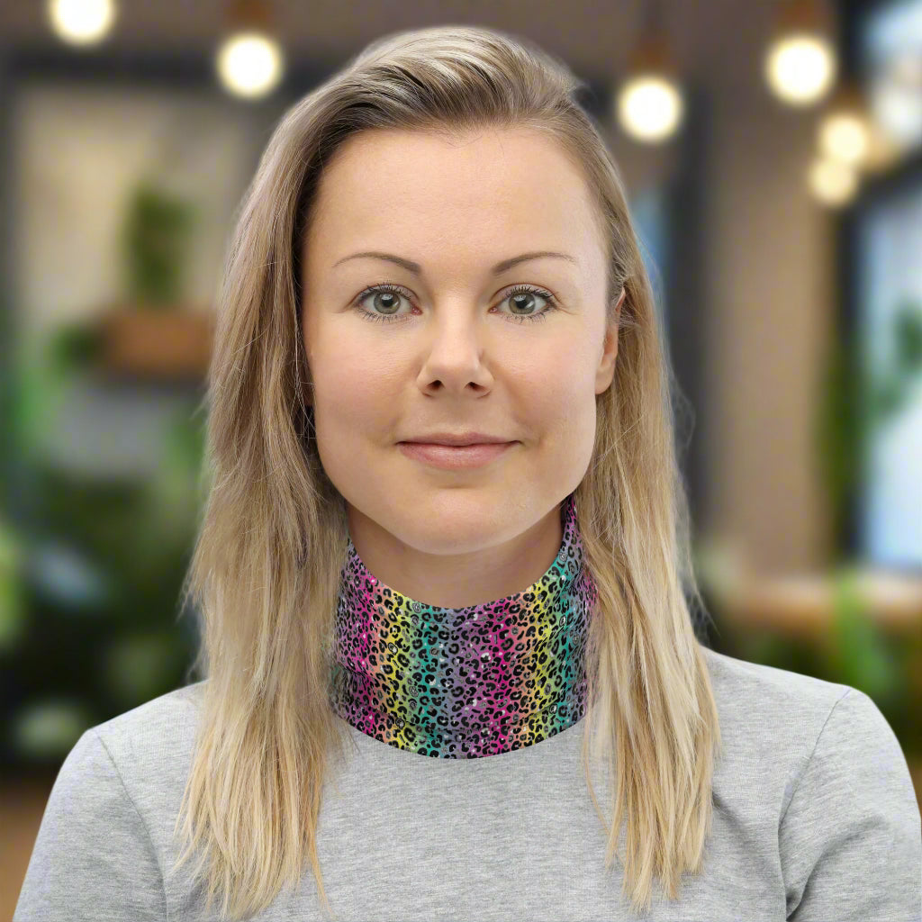 Woman with long blonde hair facing to the front wearing a grey long sleeve top and a vibrant rainbow leopard glamour neck gaiter around her neck. In the background are some cafe tables and plants. The rainbow leopard glamour design is an animal print pattern in black, grey and white, printed at a small scale against a blended rainbow background