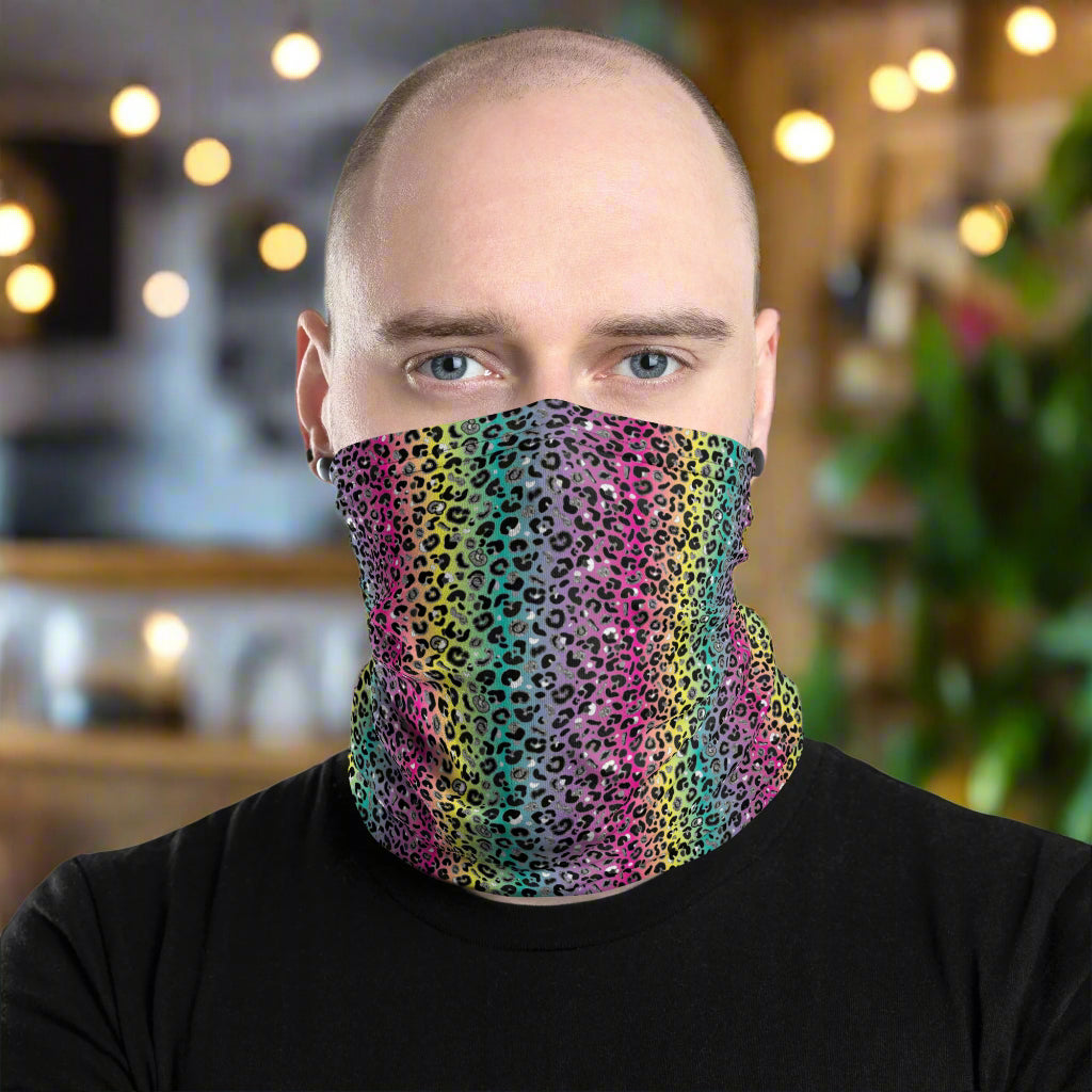 Bald man facing to the front wearing a black top and a vibrant rainbow leopard glamour neck gaiter around his neck, covering his mouth and nose. In the background are some fairy lights and plants. The rainbow leopard glamour design is an animal print pattern in black, grey and white, printed at a small scale against a blended rainbow background