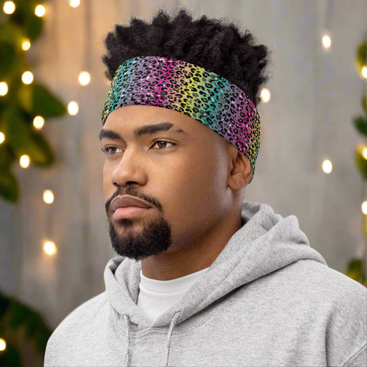 Man with short afro hair facing to the left wearing a grey top and a vibrant rainbow leopard glamour neck gaiter around his head as a head band. In the background are some fairy lights and plants. The rainbow leopard glamour design is an animal print pattern in black, grey and white, printed at a small scale against a blended rainbow background