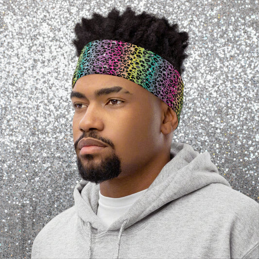 Man with short afro hair facing to the left wearing a grey top and a vibrant rainbow leopard glamour neck gaiter around his head as a head band. In the background are some fairy lights and plants. The rainbow leopard glamour design is an animal print pattern in black, grey and white, printed at a small scale against a blended rainbow background