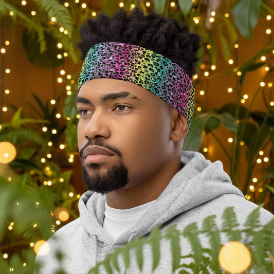 Man with short afro hair facing to the left wearing a grey top and a vibrant rainbow leopard glamour neck gaiter around his head as a head band. In the background are some fairy lights and plants. The rainbow leopard glamour design is an animal print pattern in black, grey and white, printed at a small scale against a blended rainbow background