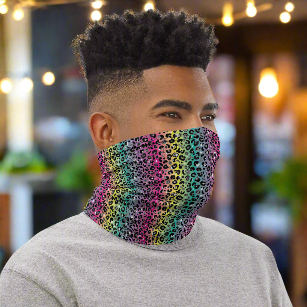 Man with short afro hair facing to the right wearing a grey top and a vibrant rainbow leopard glamour neck gaiter around his neck, covering his mouth and nose. In the background are some fairy lights and plants. The rainbow leopard glamour design is an animal print pattern in black, grey and white, printed at a small scale against a blended rainbow background